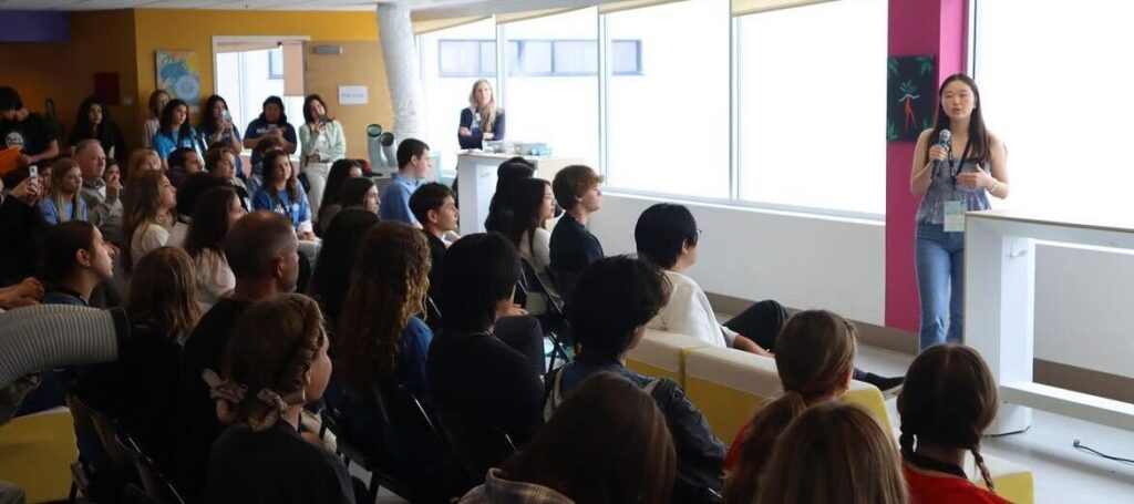 A young woman stands giving a presentation to a seated audience in a brightly lit room.