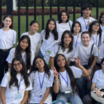 A group of high school-aged young adults, wearing matching white t-shirts and lanyards, poses for a photo in front of the White House in Washington, DC.