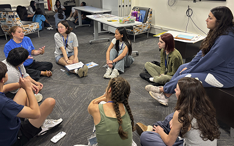 A group of young people, some wearing lanyards, sit on a gray carpet in a circle, attentively listening to a woman in a blue shirt who is speaking and gesturing with her hands.